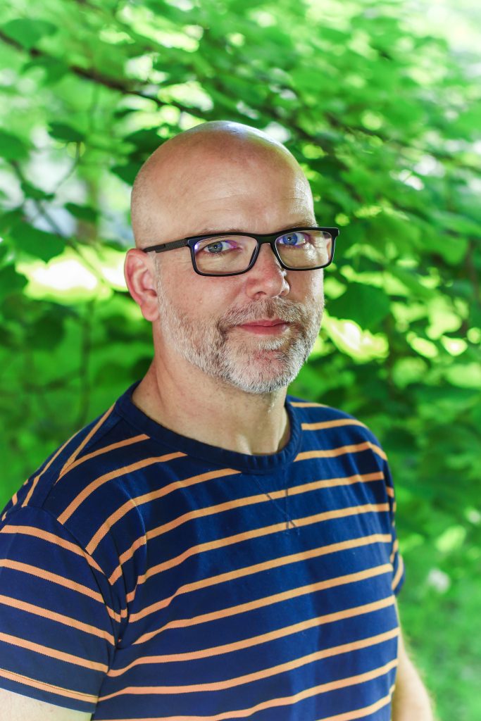 Joshua Wilbur standing outside in front of a tree with green foliage.